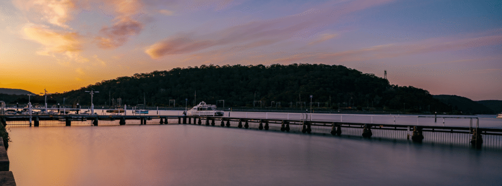 Sunset over the Central Coast waterfront with calm waters, boats docked at the marina, and a tree-covered hill in the background.